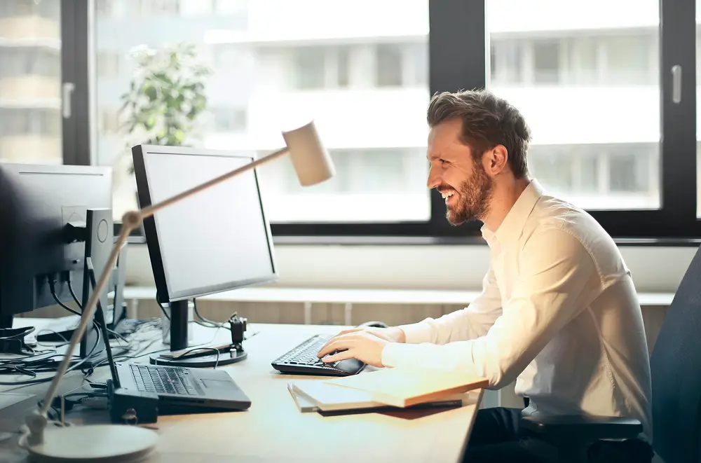 Ein Mann im weißen Hemd arbeitet lächelnd an einem Schreibtisch mit einem Computer in einem hellen Büro und plant das Eventmanagement für einen Freizeitpark. Große Fenster und eine Schreibtischlampe sind in der Nähe.