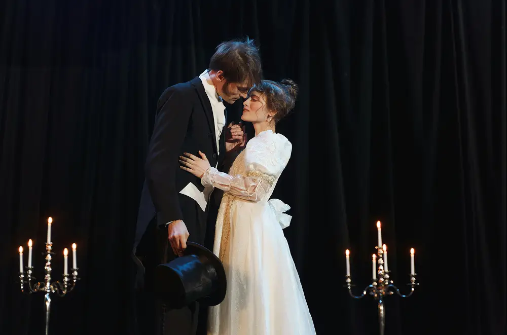 Ein Mann im Anzug und eine Frau in einem weißen Vintage-Kleid stehen dicht beieinander auf der Bühne, umgeben von beleuchteten Kerzenleuchtern und schwarzen Vorhängen. Die dramatische Theaterkulisse wird noch verstärkt, da der Mann einen Zylinder hält.