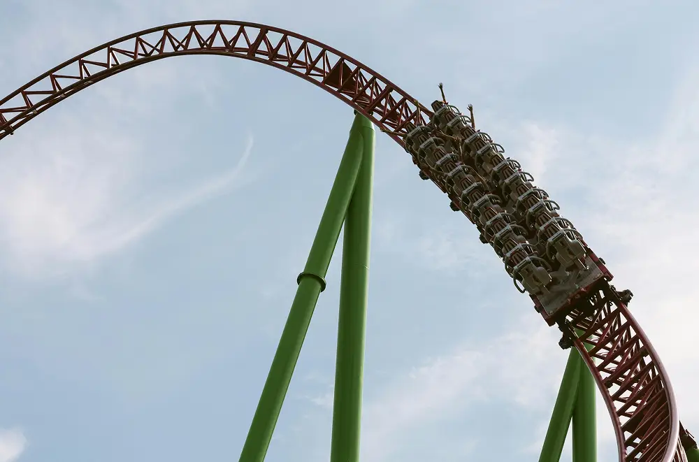 Ein Achterbahnzug mit Fahrgästen fährt eine steile, rote Strecke hinauf, die von hohen grünen Balken vor einem blauen Himmel gestützt wird - ein aufregendes Highlight in jeder Eventmanagement-Strategie für ein lebendiges Freizeitpark-Erlebnis.