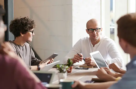Vier Personen sitzen um einen Tisch in einem hellen Büro und halten eine Besprechung ab. Sie verwenden digitale Tablets und Laptops, eine Person hält einen Stift und spricht, während die anderen zuhören und Notizen machen. Eine kleine Pflanze steht auf dem Tisch.