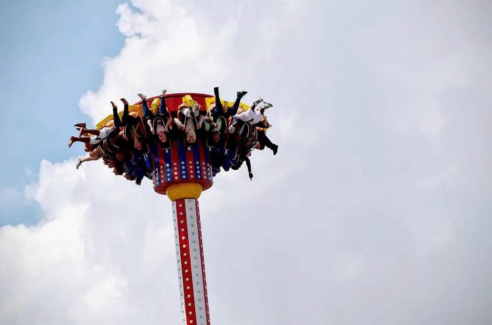 Eine Gruppe von Menschen auf einer Fallturmfahrt in einem Vergnügungspark, die mit baumelnden Beinen hoch in der Luft schwebt, fängt den Nervenkitzel und die Koordination ein, die man oft im digitalen Eventmanagement findet, und das alles vor dem Hintergrund eines bewölkten Himmels.