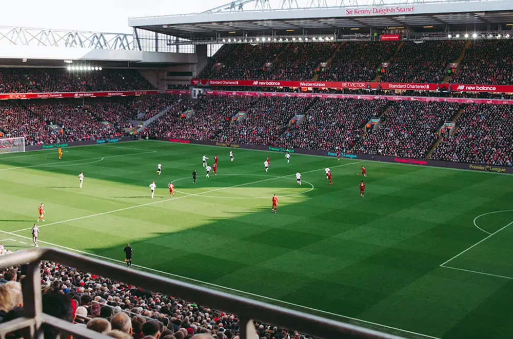 Ein volles Fußballstadion zeigt Eventmanagement Sport auf höchstem Niveau, während die Fans ein spannendes Spiel zwischen den Mannschaften in Rot und Weiß auf einem leuchtend grünen Spielfeld unter einem klaren Himmel verfolgen.