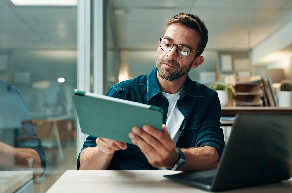 Ein Mann mit Brille sitzt an einem Schreibtisch mit einem Laptop und schaut auf ein Tablet-Gerät in seinen Händen. Er scheint konzentriert zu sein, und der Arbeitsbereich hat eine moderne, professionelle Atmosphäre mit Regalen und Pflanzen im Hintergrund.