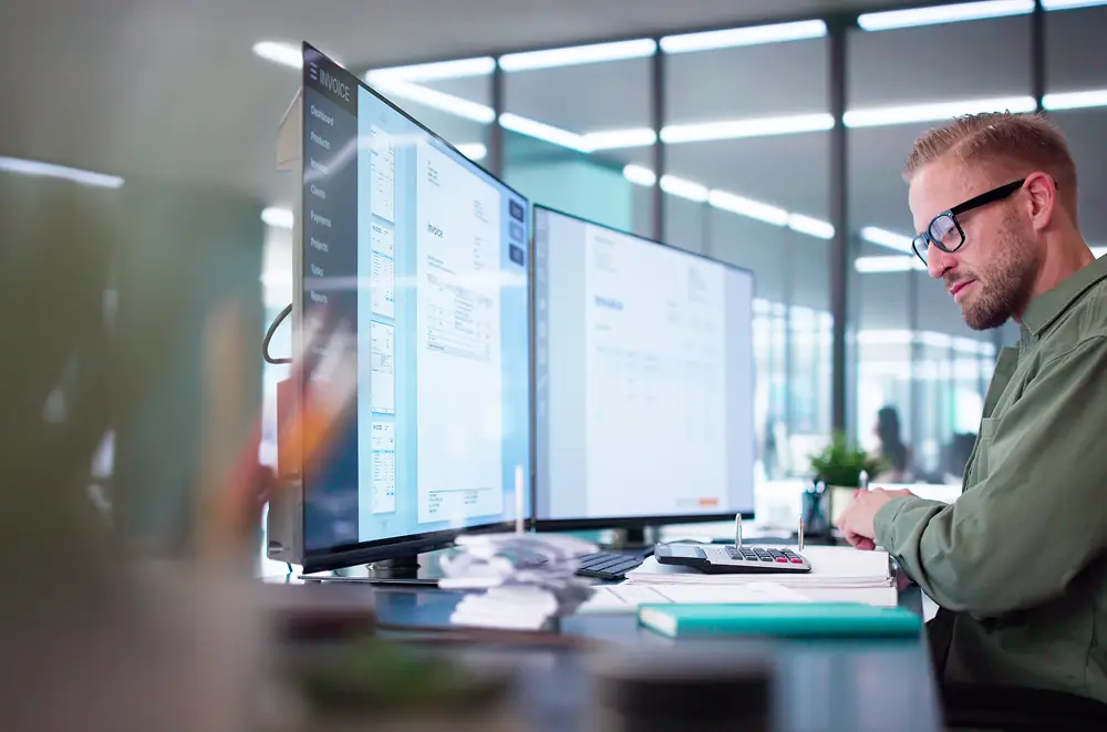 Ein Mann mit Brille arbeitet an einem Schreibtisch mit zwei großen Computermonitoren, einem Taschenrechner und Papieren in einem modernen Büro mit Glaswänden und heller Beleuchtung und setzt die Digitale Transformation in seinem täglichen Arbeitsablauf um.