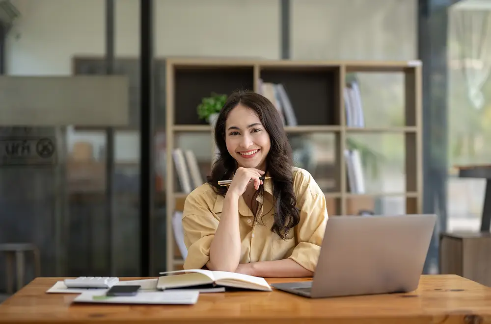 Eine Frau mit langen dunklen Haaren sitzt an einem Schreibtisch mit einem Laptop, einem Notebook und Eventmanagement-Zoopapieren und lächelt in die Kamera in einem modernen Büro mit Bücherregalen im Hintergrund.