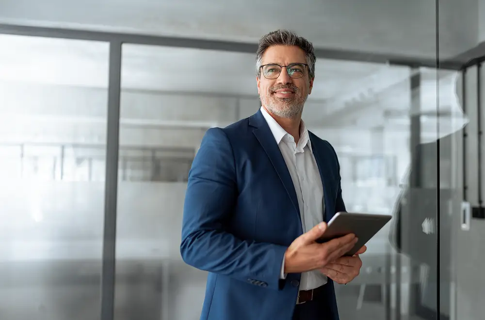 Ein selbstbewusster Mann mittleren Alters im blauen Anzug und mit Brille steht in einem modernen Büro, lächelt und hält ein Tablet in der Hand - ein Symbol der digitalen Transformation. Glaswände und helle Beleuchtung schaffen ein professionelles Geschäftsumfeld.