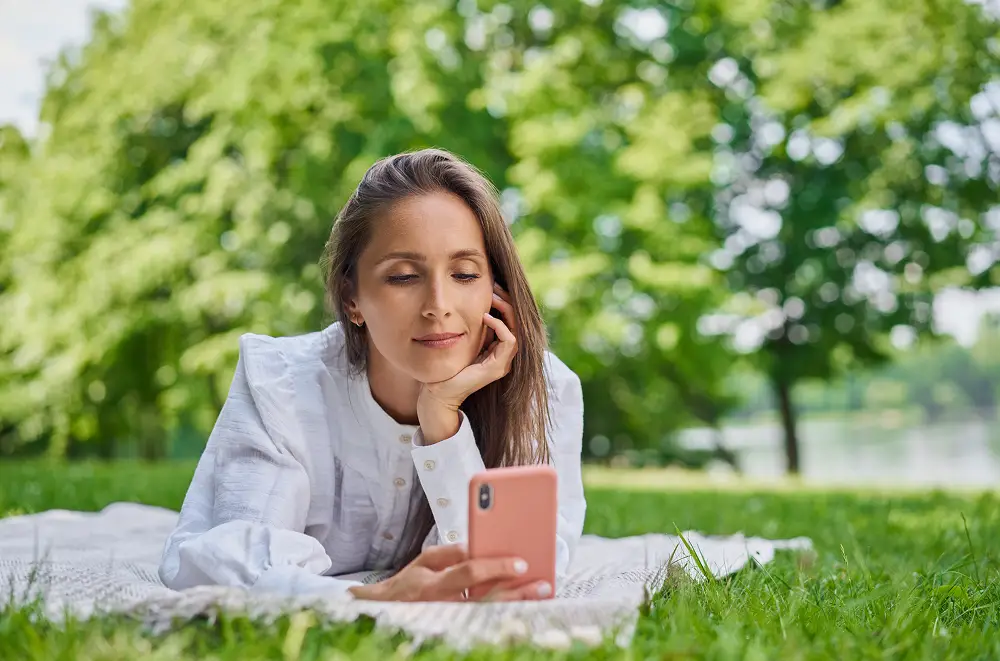 Eine Frau in einem weißen Hemd liegt auf einer Decke im Gras, schaut auf ihr Smartphone und lächelt, während hinter ihr grüne Bäume und ein unscharfer Outdoor-Hintergrund zu sehen sind.