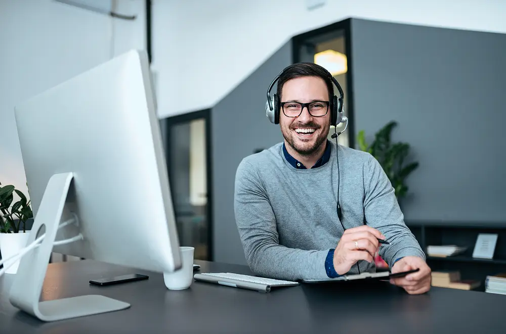Ein lächelnder Mann mit Brille und Headset sitzt an einem Schreibtisch mit Computermonitor, Tastatur und Notizblock in einer modernen Büroumgebung.