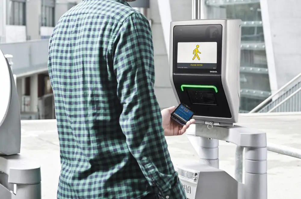 Eine Person in einem grün karierten Hemd hält ein Smartphone in die Nähe eines elektronischen Zugangsterminals im Freien, das ein Symbol für eine gehende Person und die Worte ZUGANG GEWÄHRT auf seinem Bildschirm anzeigt.