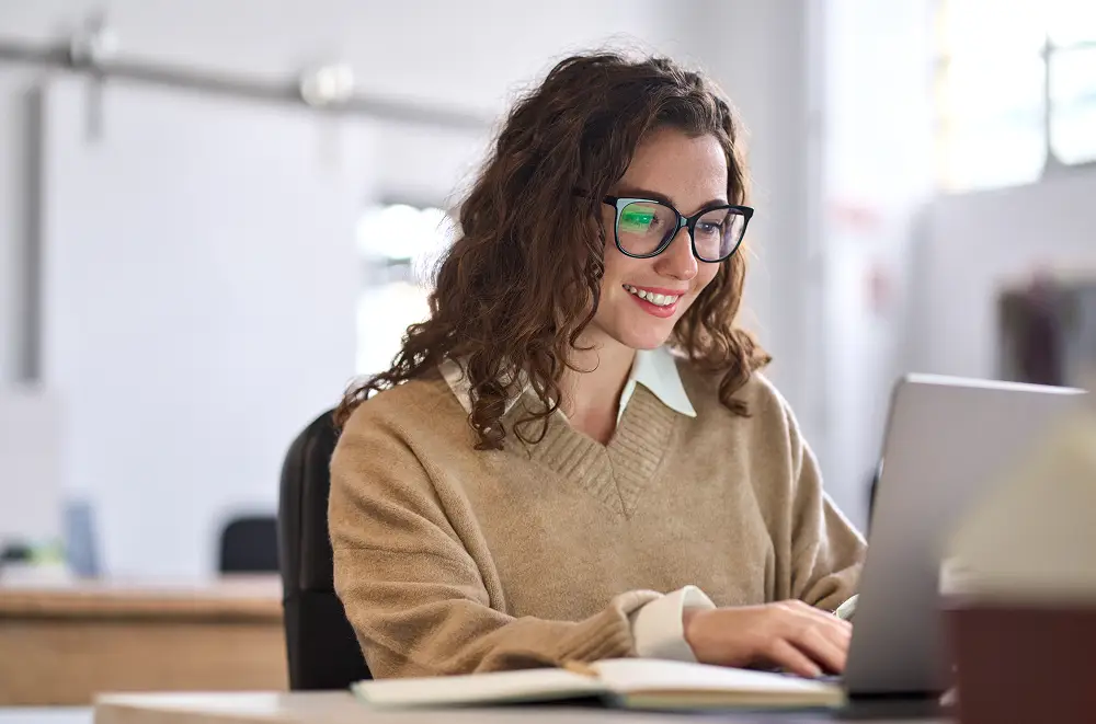 Eine Frau mit lockigem braunem Haar und Brille lächelt, während sie an einem Schreibtisch in einem hellen Büro in der Immobilienverwaltung auf einem Laptop tippt. Sie trägt einen beigen Pullover über einem Kragenhemd.