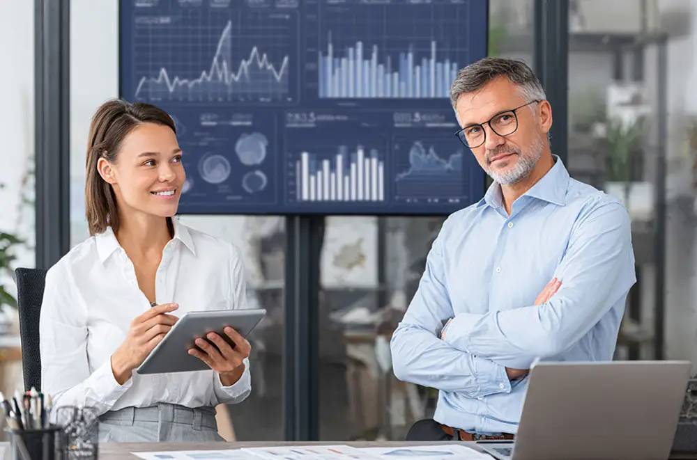 Eine lächelnde Frau, die ein Tablet in der Hand hält, und ein Mann mit verschränkten Armen stehen in einem Büro. Hinter ihnen zeigen große digitale Bildschirme Microsoft Dynamics-Diagramme und -Grafiken an. Auf dem Schreibtisch davor liegen Papiere und ein Laptop.