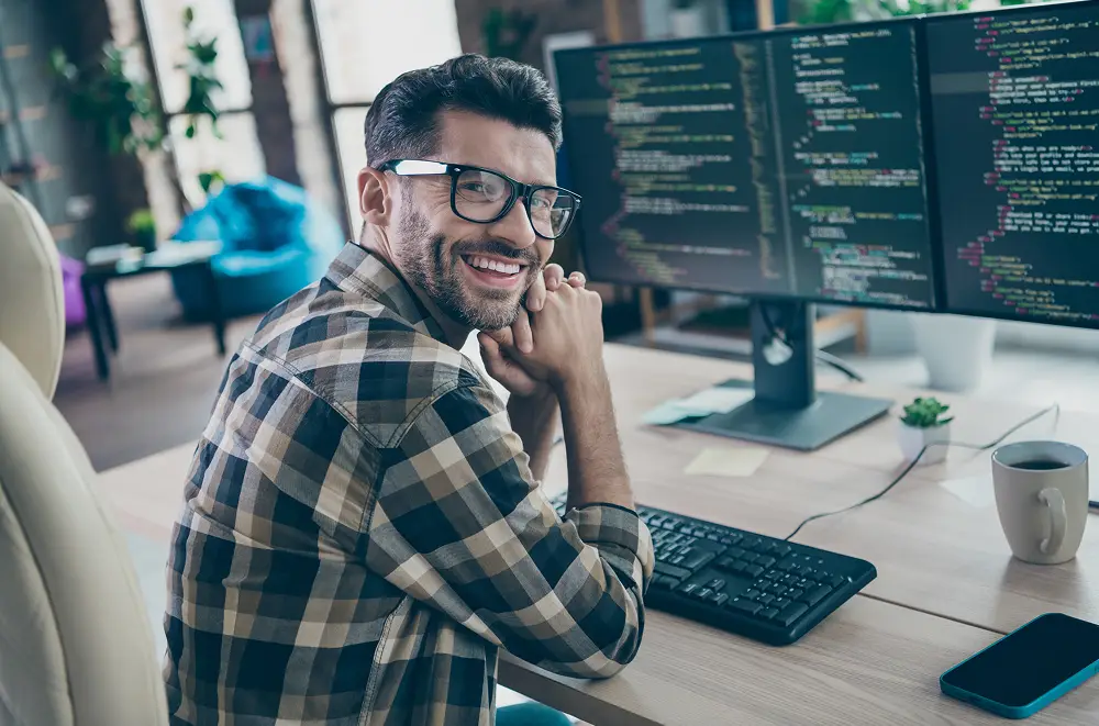 Ein lächelnder Mann mit Brille und kariertem Hemd sitzt an einem Schreibtisch mit einer Tastatur und blickt in die Kamera. Vor ihm zeigen Computermonitore Codezeilen für ein System der Immobilienverwaltung in einer modernen, hellen Büroumgebung.