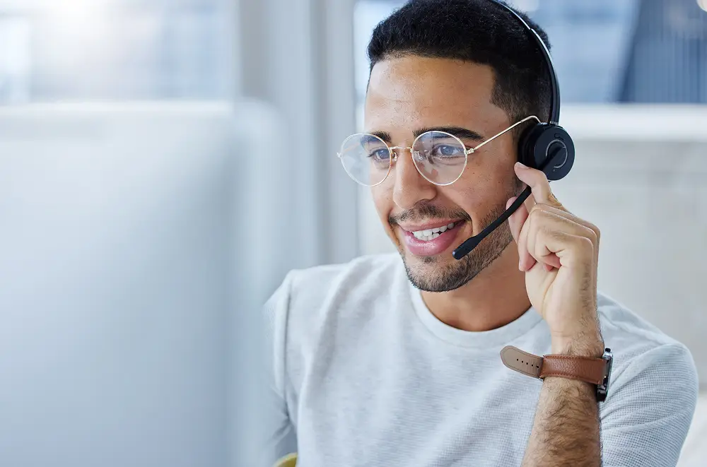 Ein lächelnder Mann mit Brille und Headset sitzt an einem Schreibtisch, schaut auf einen Computerbildschirm und berührt das Mikrofon des Headsets - bereit, Kunden im Zeitalter der Digitalisierung im Handel zu unterstützen.