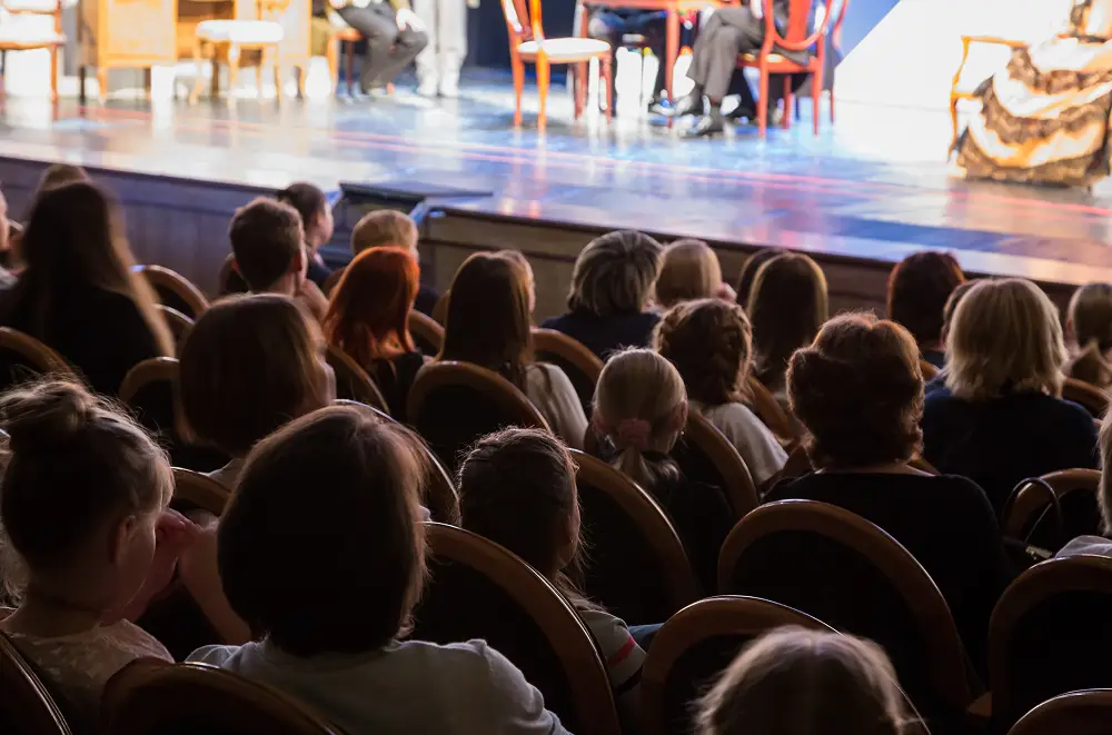Die Zuschauer sitzen in Sitzreihen und verfolgen eine Live-Theateraufführung. Die Bühne ist hell erleuchtet, Schauspieler und Requisiten sind zu sehen, während das Publikum in gedämpftem Licht sitzt - ein Zeichen für fachkundiges Eventmanagement.