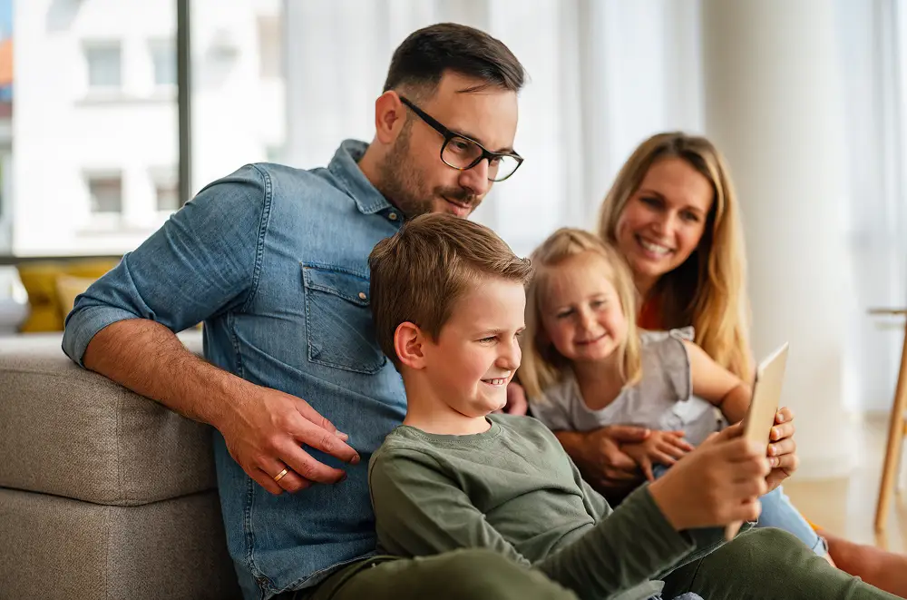 Eine lächelnde vierköpfige Familie sitzt zusammen auf einem Sofa, zwei Kinder halten ein Tablet in der Hand, die Eltern dicht dahinter. Alle schauen glücklich auf den Bildschirm, während sie in ihrem hellen Wohnzimmer eine Eventmanagement-Zoo-Tour erkunden.
